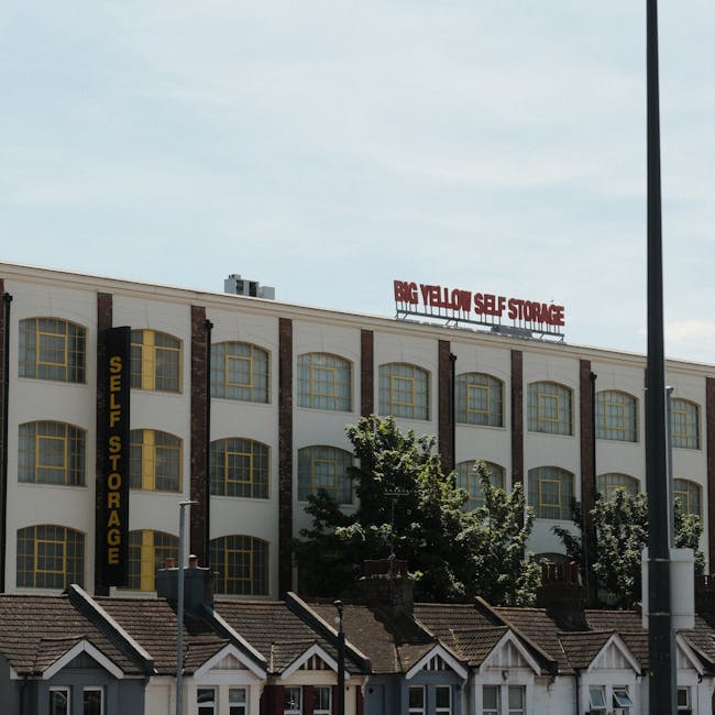 Photograph of a large, multi-storey building with a white facade and brick accents, featuring multiple rectangular windows with yellow frames. A vertical sign on the building reads 'SELF STORAGE', and a red sign on the roof displays 'BIG YELLOW SELF STORAGE' in capital letters. In front of the building, there are smaller residential houses with pitched roofs, chimney stacks, and small windows, partially obscured by a row of trees with dense foliage. The scene is set outdoors during daylight hours, with a clear sky and sunlight illuminating the structures. A tall street lamp is visible on the right side of the image, and the ground in front of the houses appears to be a pavement area. The image depicts an urban environment suitable for house removals and furniture transport, with the self-storage facilities providing additional options for packing and moving logistics, as handled by companies like Man With a Van Homerton.