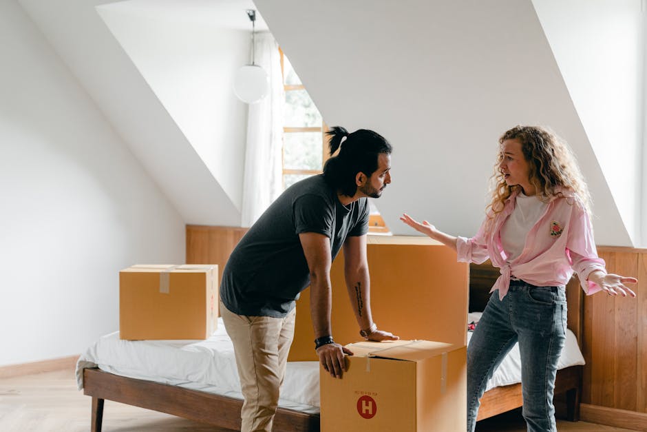 A man with dark hair tied back, wearing a dark t-shirt and beige trousers, is inside a room with sloped ceiling and wooden paneling along one wall, engaged in a conversation with a woman who has curly blonde hair and is dressed in a pink top and jeans. They are surrounded by moving boxes of various sizes, some open with packing materials, and placed on and around a bed with white bedding. The man is leaning on a box, and the woman is gesturing with her hands, appearing to discuss the packing or home relocation process. The room is well-lit with natural daylight coming through a window, and a white pendant light hangs from the ceiling. The scene captures a typical furniture transport and packing stage of house removals, illustrating the planning and coordination involved in professional moving services by [COMPANY_NAME].