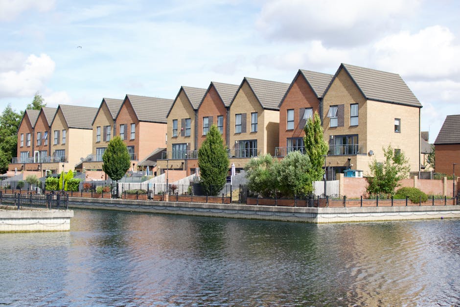 Photograph of a large, multi-storey building with a white facade and brick accents, featuring multiple rectangular windows with yellow frames. A vertical sign on the building reads 'SELF STORAGE', and a red sign on the roof displays 'BIG YELLOW SELF STORAGE' in capital letters. In front of the building, there are smaller residential houses with pitched roofs, chimney stacks, and small windows, partially obscured by a row of trees with dense foliage. The scene is set outdoors during daylight hours, with a clear sky and sunlight illuminating the structures. A tall street lamp is visible on the right side of the image, and the ground in front of the houses appears to be a pavement area. The image depicts an urban environment suitable for house removals and furniture transport, with the self-storage facilities providing additional options for packing and moving logistics, as handled by companies like Man With a Van Homerton.