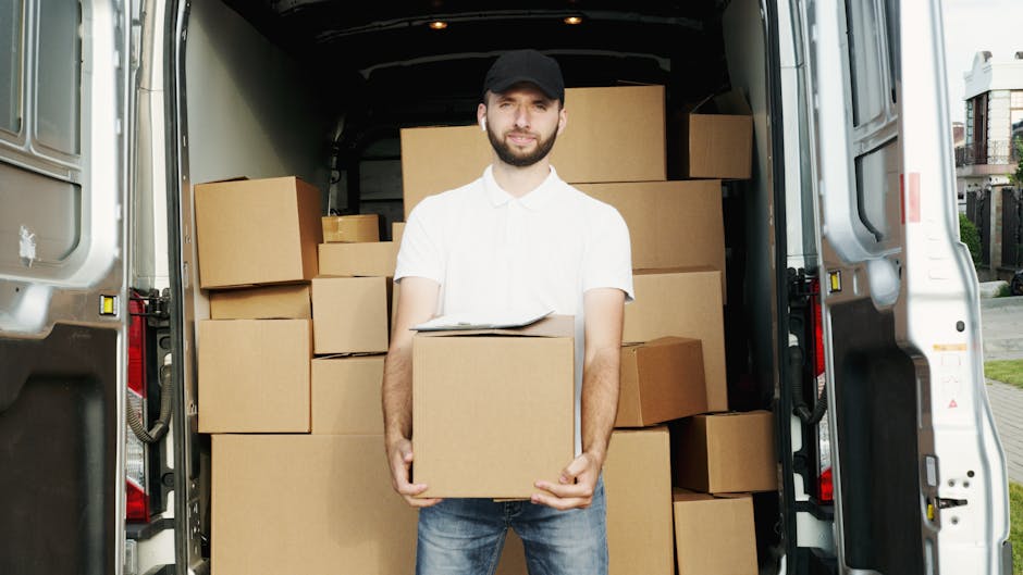 A man wearing a blue jacket and dark trousers is seated on the back of an open, white delivery van with visible wooden paneling inside. He is reviewing a clipboard and holding a pen, surrounded by pac