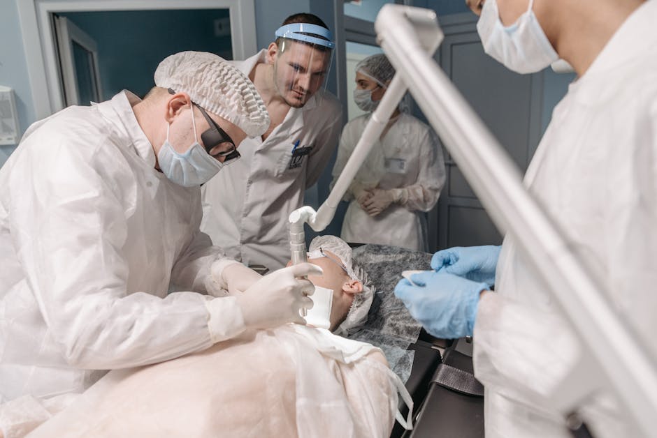 In a hospital operating theatre, a team of medical professionals, including a surgeon wearing glasses, a surgical cap, and a surgical mask, is performing a procedure on a patient lying on the operatin