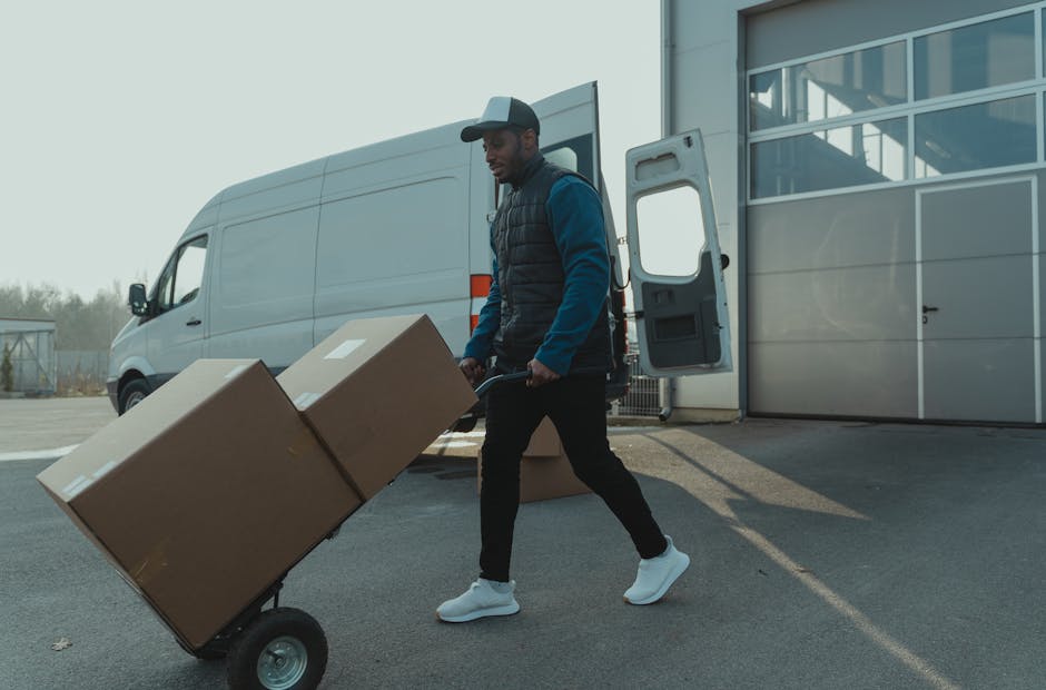 A man wearing a baseball cap, dark trousers, a blue jacket, and white sneakers is pushing a hand truck loaded with two large cardboard boxes outside a modern commercial building with glass panels. The loading area is paved, and a large white delivery van with its rear doors open is parked nearby, indicating a home relocation or furniture transport process. The van's interior and the open doors reveal packaging materials like plastic wrap and blankets used for safe packing in house removals. The scene is lit with natural daylight, highlighting the careful handling of household items during packing and moving activities, with [COMPANY_NAME] providing removal services in the context of a same-day move, as referenced on the Homerton removal guide webpage.