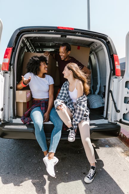 A smiling man and two women sitting inside the open rear of a large moving van, with visible cardboard boxes and wrapped furniture around them, on a paved driveway near a home. One woman has curly hair and is wearing a white T-shirt and blue jeans, the other woman has long straight hair and is dressed in a white top with a checkered shirt tied around her waist, while the man stands between them, all engaged in conversation. The van's interior shows various packing materials such as plastic wrap and blankets, and the back doors are open to facilitate the loading process. The scene depicts a home relocation moment during packing and moving activities, with clear natural daylight illuminating the scene, highlighting the collaborative effort involved in furniture transport and household packing for a remova service by Man With a Van Homerton.