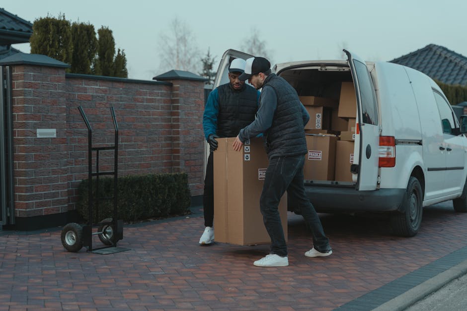 A man wearing a baseball cap, dark trousers, a blue jacket, and white sneakers is pushing a hand truck loaded with two large cardboard boxes outside a modern commercial building with glass panels. The loading area is paved, and a large white delivery van with its rear doors open is parked nearby, indicating a home relocation or furniture transport process. The van's interior and the open doors reveal packaging materials like plastic wrap and blankets used for safe packing in house removals. The scene is lit with natural daylight, highlighting the careful handling of household items during packing and moving activities, with [COMPANY_NAME] providing removal services in the context of a same-day move, as referenced on the Homerton removal guide webpage.