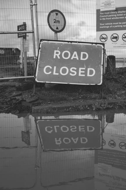 A black and white photograph showing a large rectangular 'Road Closed' sign leaning against a metal barrier at a construction site or roadwork area, with muddy ground and scattered debris in the foreg